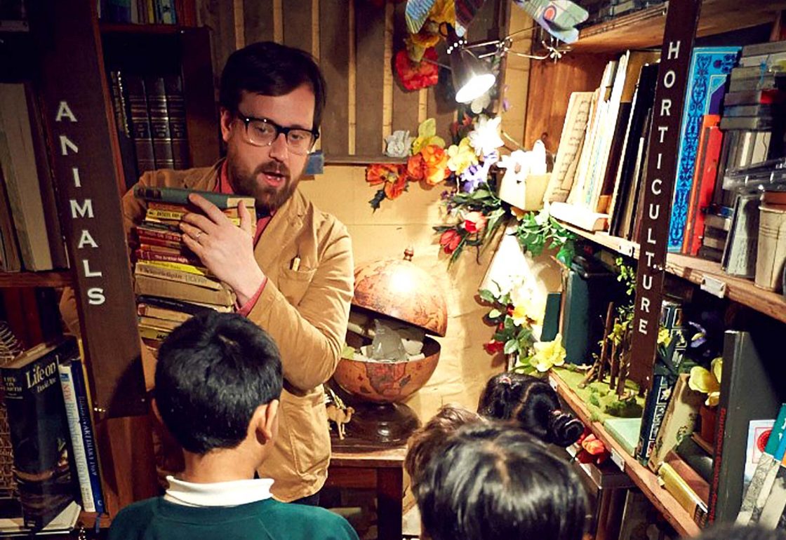 An image of a teacher holding a stack of books with pupils participating in a Punchdrunk theatrical experiences, a charity supported by the Ellis Campbell Foundation, helping disadvantaged young people in Hampshire, London and Perthshire