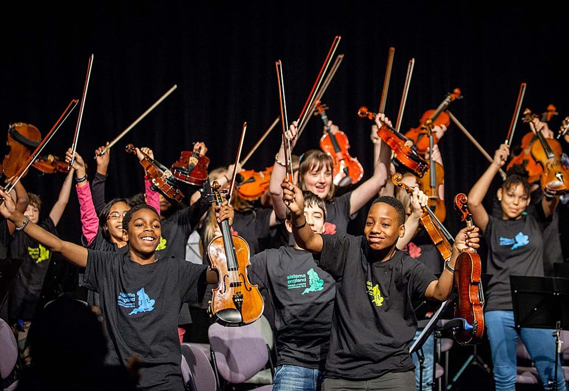 An image of pupils with violins and bows held in the air, taking part in a social action music programme run by Sistema, a charity supported by the Ellis Campbell Foundation, helping disadvantaged young people in Hampshire, London and Perthshire
