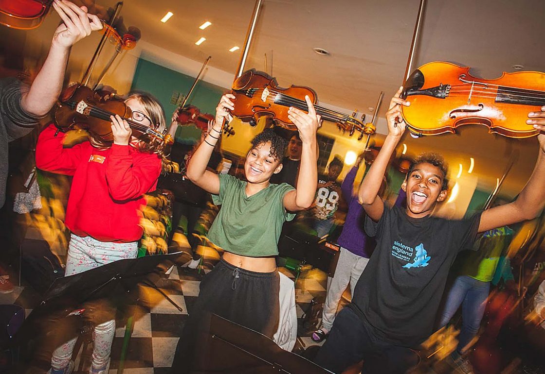 An image of pupils with violins and bows held in the air, taking part in a social action music programme run by Sistema, a charity supported by the Ellis Campbell Foundation, helping disadvantaged young people in Hampshire, London and Perthshire