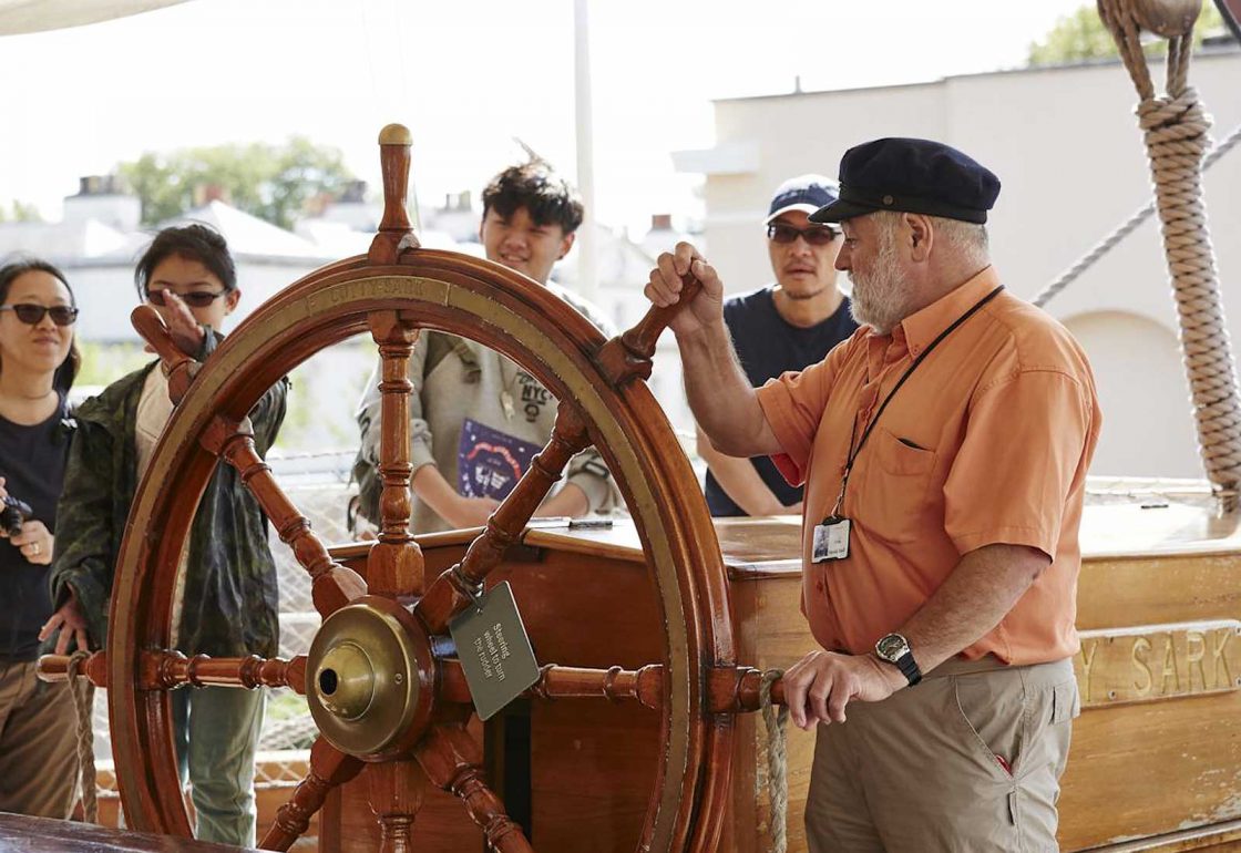 An image of a man at the helm of the Cutty Sark with onlookers representing the Lifeboats Conservation Project grant at the Royal Museum Greenwich made by the Ellis Campbell Foundation helping disadvantaged young people in Hampshire, London and Perthshire