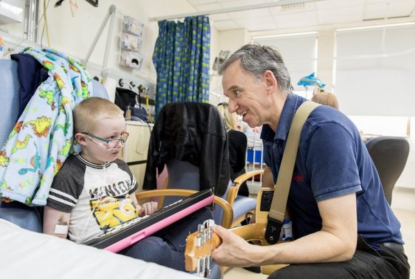 A colour image of a man playing a guitar to a sick child in hospital - part of Young Sparks supported by the Kensington & Chelsea Foundation - A grant made by The Ellis Campbell Foundation