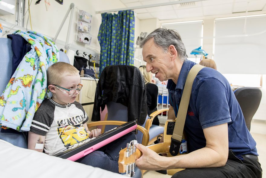 A colour image of a man playing a guitar to a sick child in hospital - part of Young Sparks supported by the Kensington & Chelsea Foundation - A grant made by The Ellis Campbell Foundation