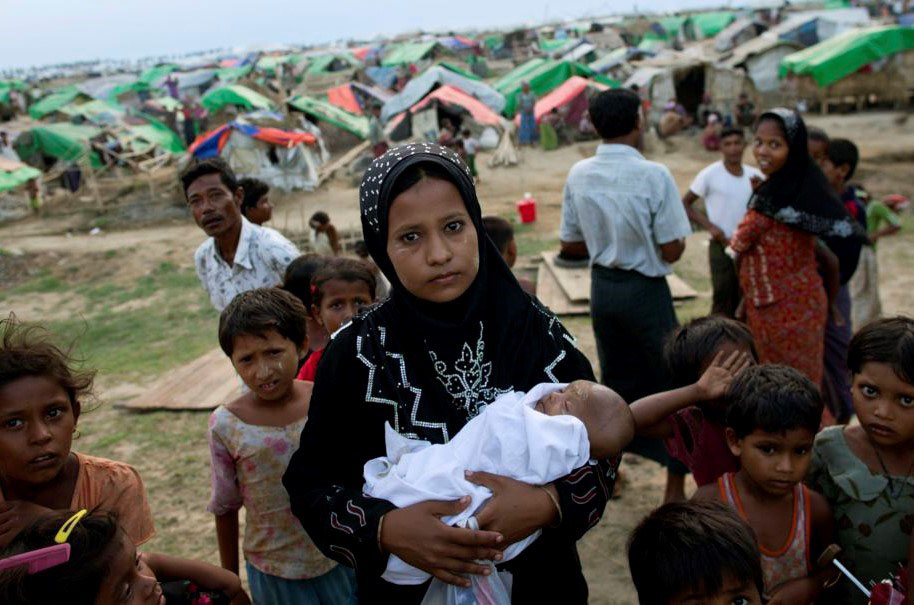 A colour image of a displaced Rohingya woman holding a new born baby surrounded by children in the foreground of makeshift tent camps - the DEC Myanmar Appeal is supported by the Ellis Campbell Foundation