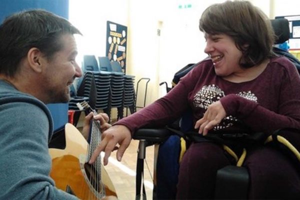 A colour image of young smiling disabled girl enjoying ma man playing a guitar as part of Parity for disability supported by the Ellis Campbell Foundation