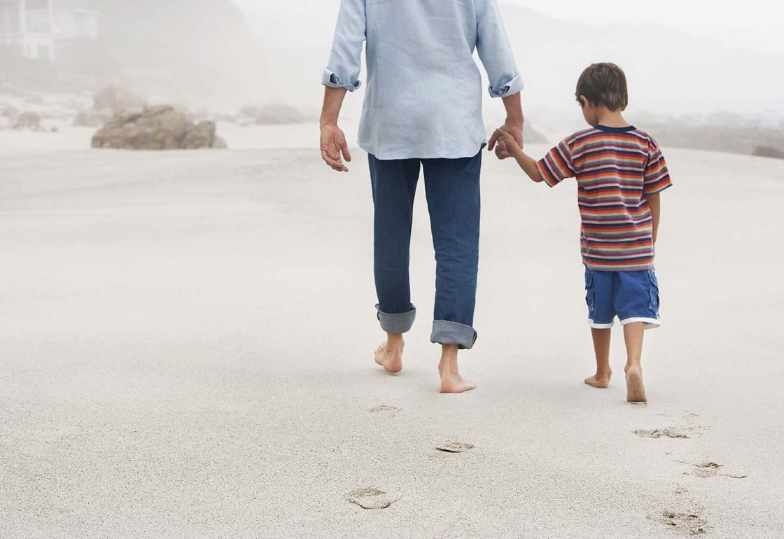 A colour picture of a caring adult holding a litle boys hand and walking along the beach - The Family Holiday Association is supported by The Ellis Campbell Foundation