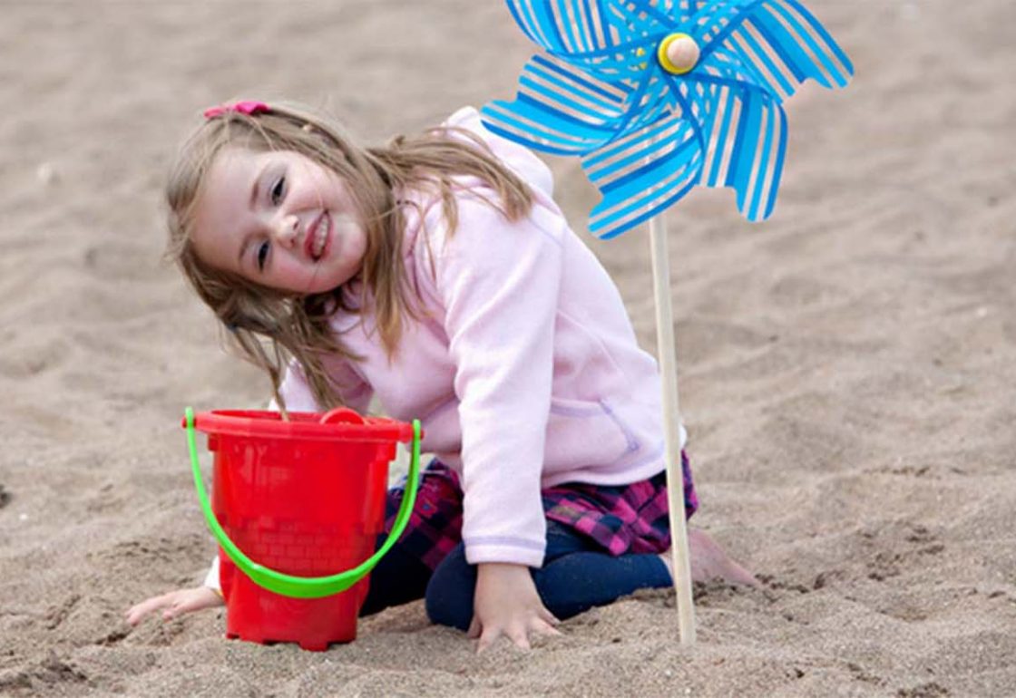A colour pciture of a little girls smiling on a beach with a bucket and blue windmill - The Family Holiday Association is supported by The Ellis Campbell Foundation