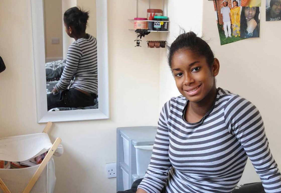 A colour image of a young black smiling female sat in her bedroom, part of the Centrepoint programme to help the homeless supported by The Ellis Campbell Foundation